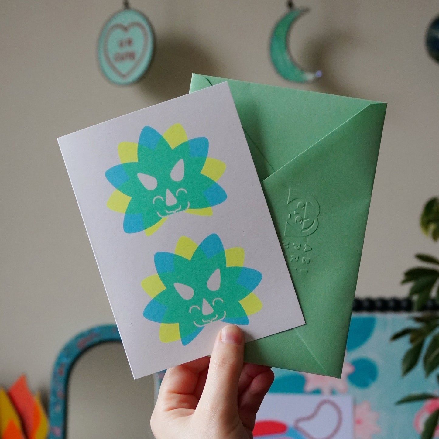 Hand holding a card with two Triceratops head designs and a green envelope against a blurred background