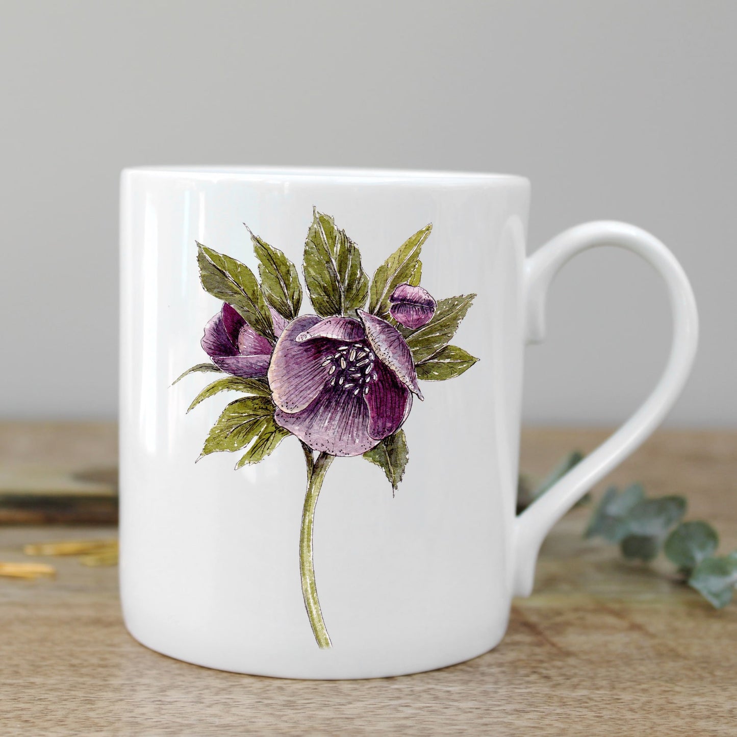 White mug with an illustration of a hellebore flower on it. Photographed on a wooden surface.
