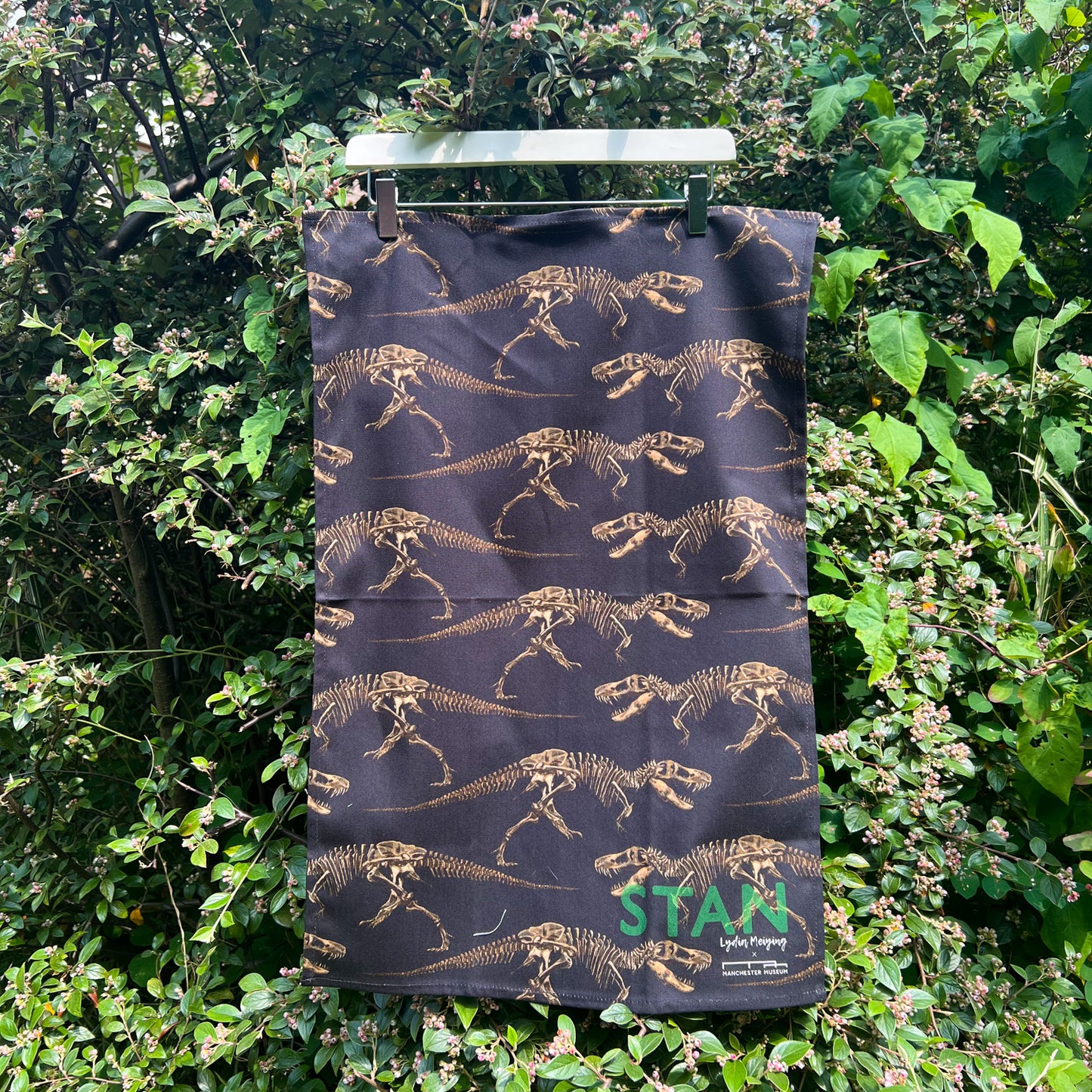Tea towel with repeat dinosaur pattern hung on a hanger in front of a garden surface
