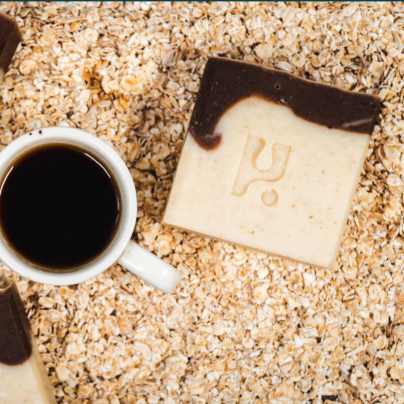 Cream and brown vegan soap bar with Och Logo imprint on a oat covered surface with a coffee next to it.