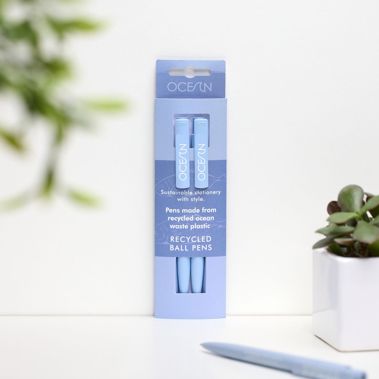 Lifestyle of a pack of two blue pens with a third blue pen in front of the pack and a small plant to the right.