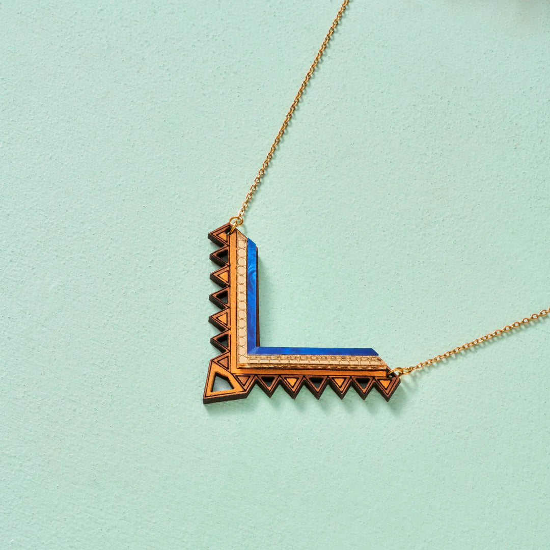 V-shaped necklace in gold and blue with triangular cutouts and pattern along the outer/bottom edge. The gold plated chain is fastened at each leg of the V. Pale sage green backdrop.