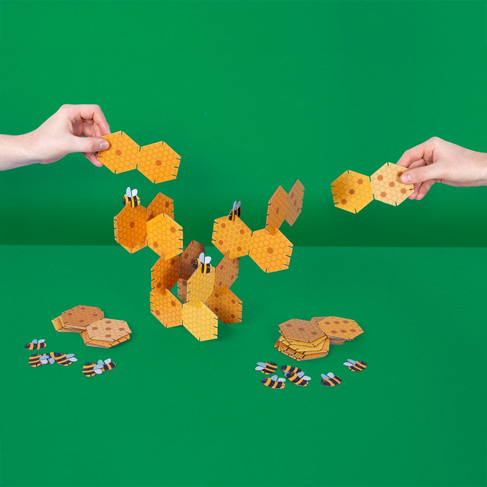 The partially assembled 3d beehive game against a green background. A hand from either side is reaching in with a puzzle piece between thumb and forefinger.