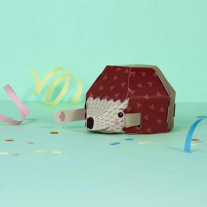 Lifestye photo of the assembled hiding hedgehog seen facing left. Teal background with colourful confetti around the hedgehog.