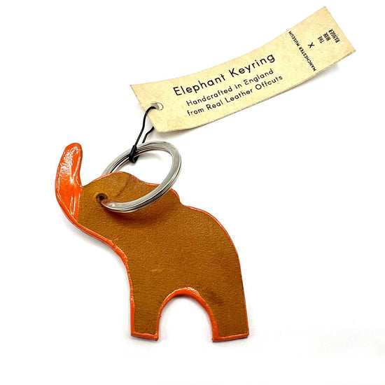 An elephant shaped keyring where the elephant stands with raised trunk. The trunk is painted orange and the same orange is painted as an outline around it's body. The keyring is where the eye would be and a paper swing tag is fastened to the keyring with a black safety pin. Tag text: Elephant keyring. Handcrafted in England from real leather offcuts.