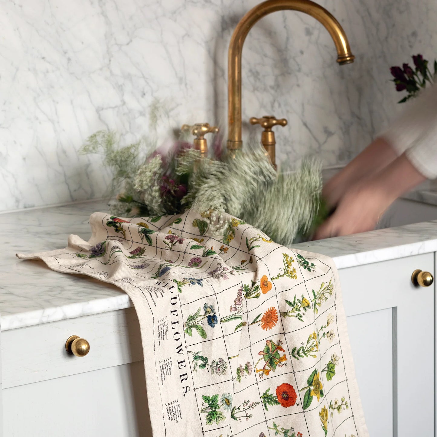 This is a close-up shot of a cotton tea towel with a grid pattern and illustrations of wildflowers, draped on a marble countertop next to a gold faucet and sink where someone is washing their hands.