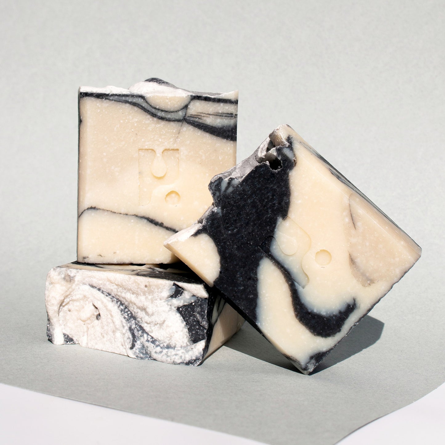 This is a shot of three square bars of soap with a swirled pattern of black and light beige colours, stacked on a light grey surface.