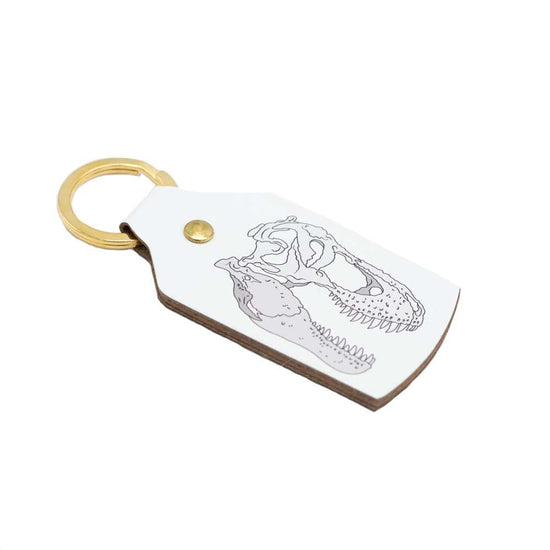The white recycled leather keyring with a t-rex skull illustration printed on the leather. The key ring is gold.