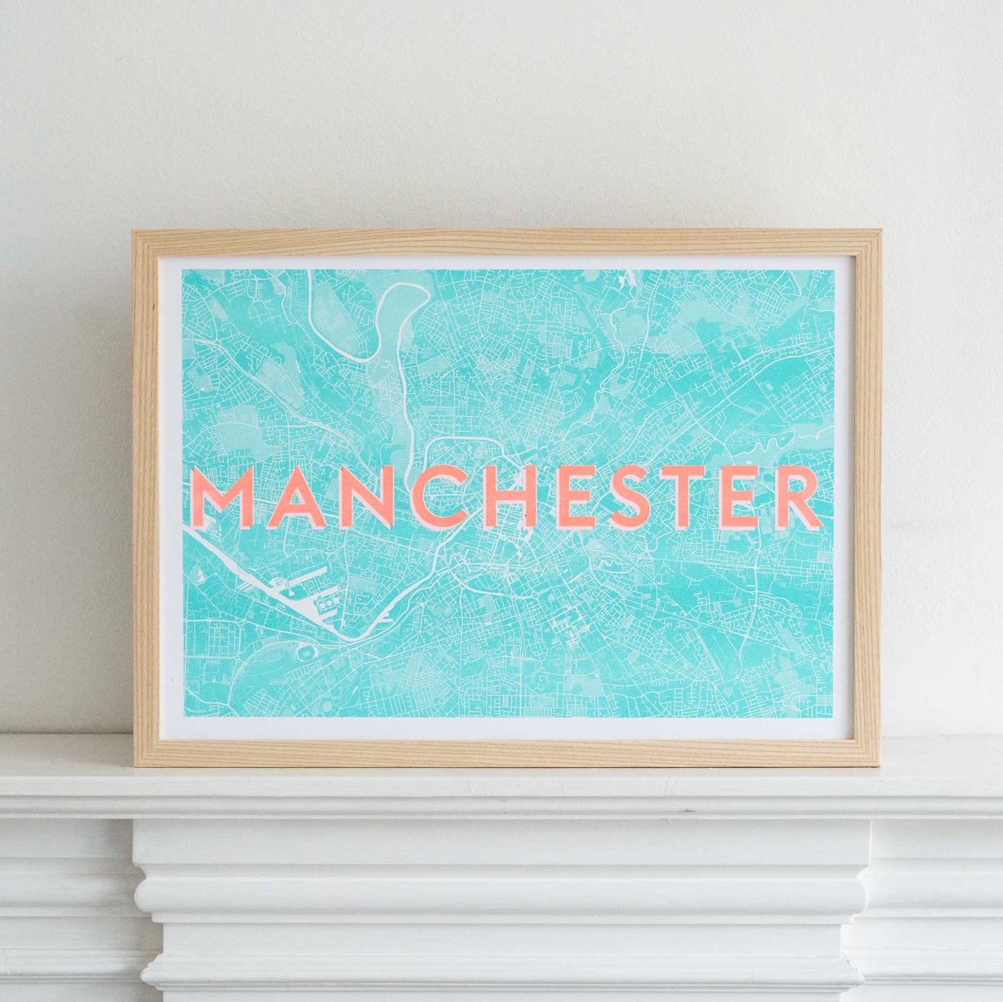 A picture of a risograph print in an ash frame. The words MANCHESTER are printed in orange above a blue and white map illustration of Manchester. Photographed on a white mantlepiece.
