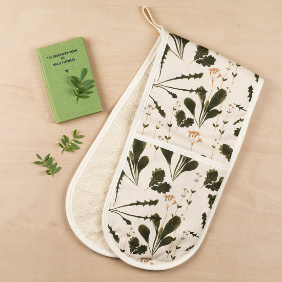 Lifestyle shot of the folded oven mittens on a wooden surface with a green book of wildflowers beside it.