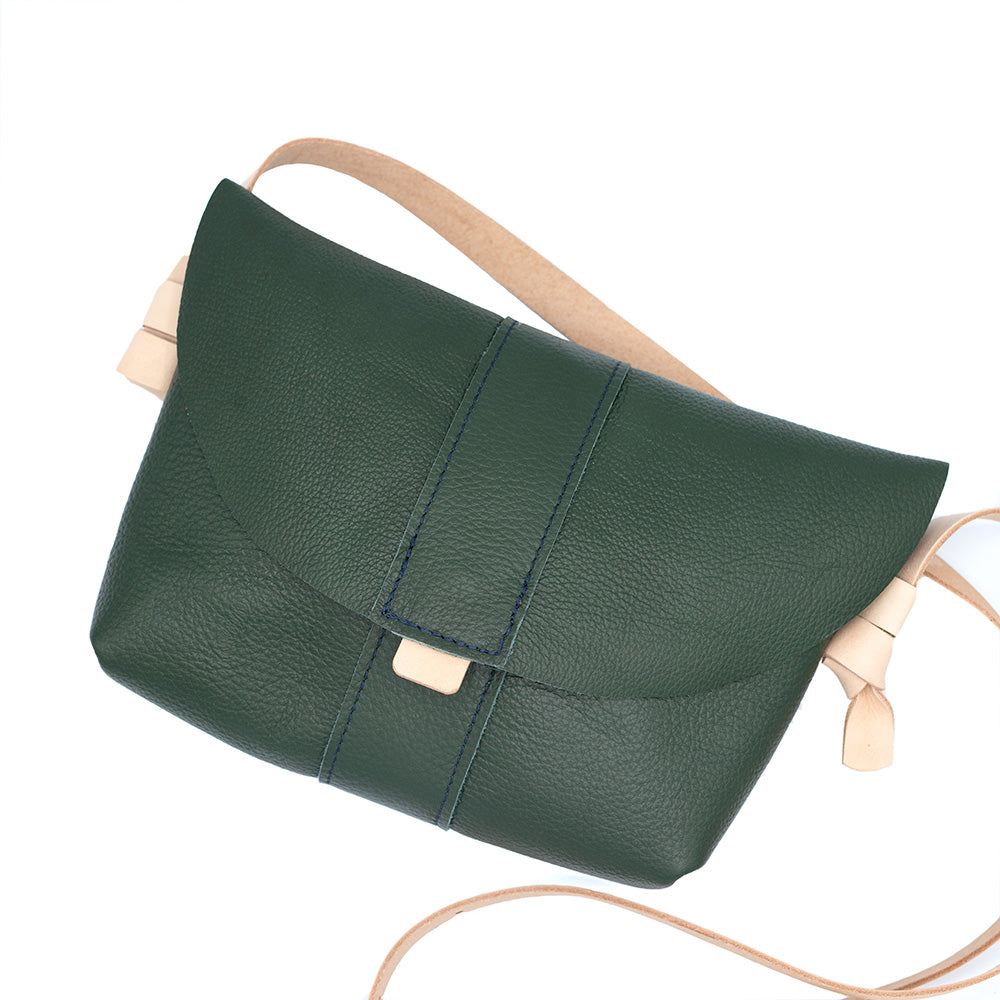 Dark green leather bag with natural pale leather strap. Seen from the front lying on a white surface.