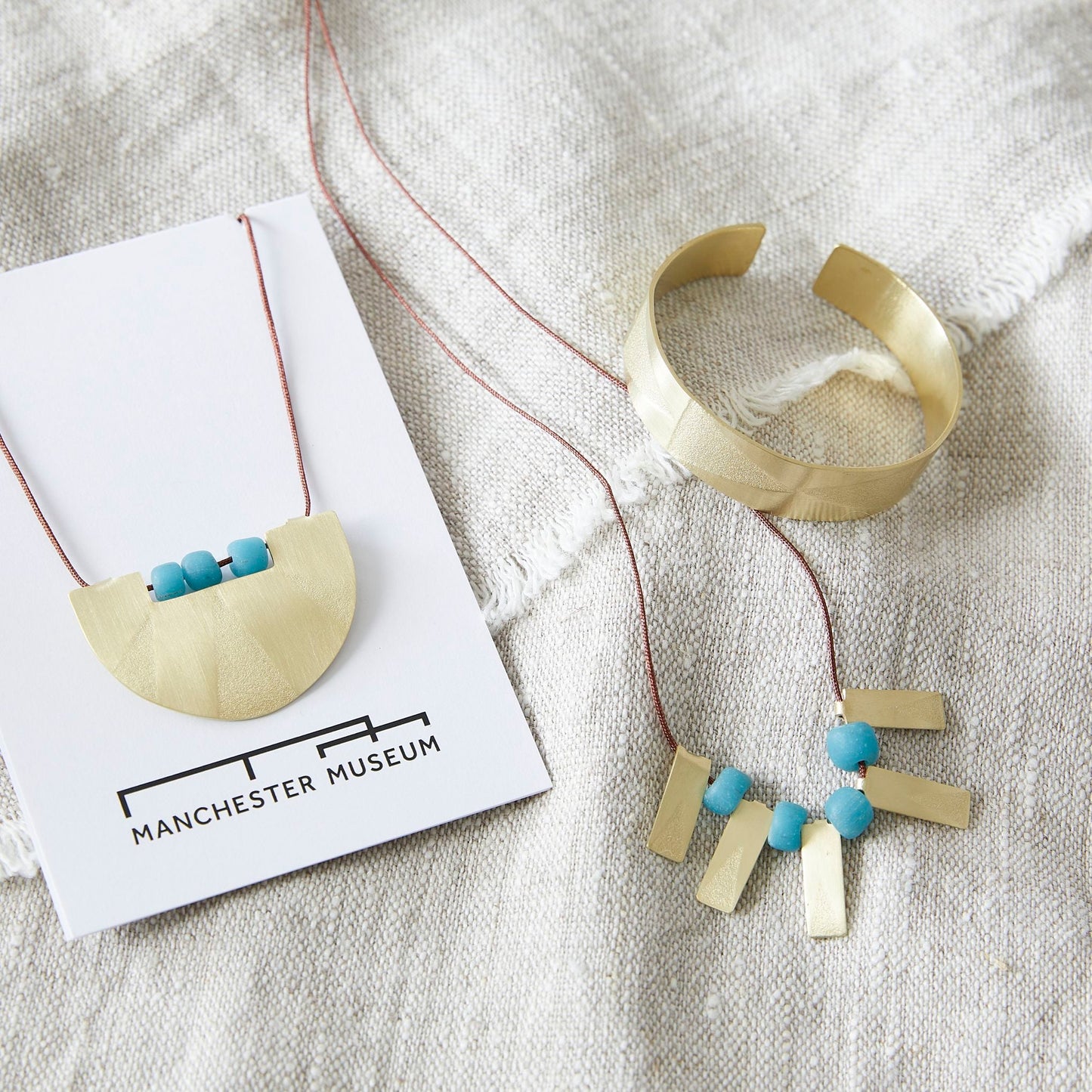 Lifestyle shot on natural linen with the semi-circular necklace to the left on the white backing card with the brass bangle and rectangular pendants necklace on the right.
