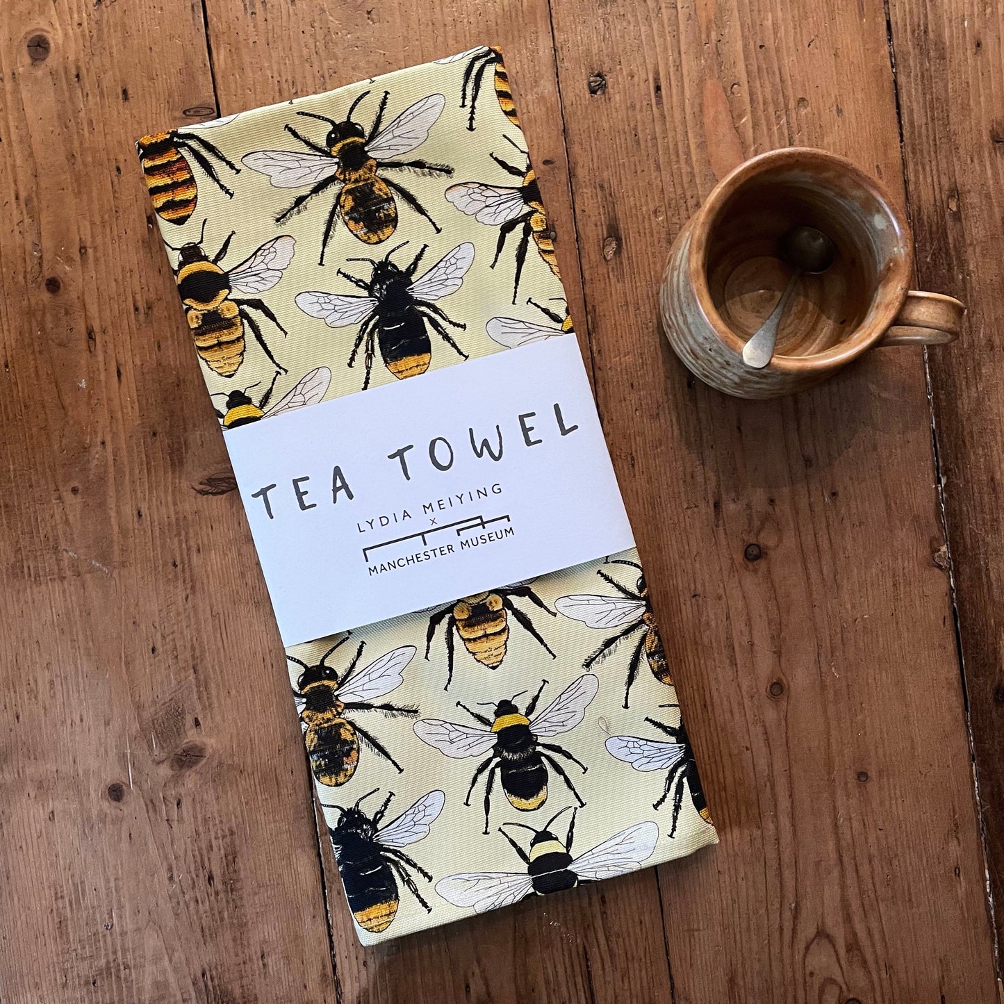 Lifestyle shot of the packaged tea towel on a rustic wooden table with an earthy coloured clay mug with a teaspoon beside it.