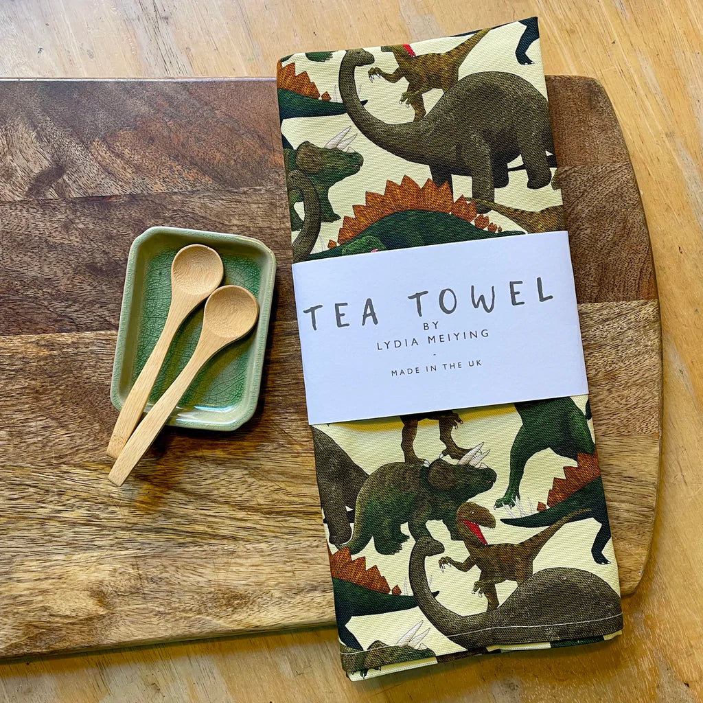 Lifestyle shot of the packaged tea towel on top of a rustic wooden chopping board. A small sage green ceramic plate is placed on th eboard beside the tea towel andtwo bamboo spoons are resting on the plate.