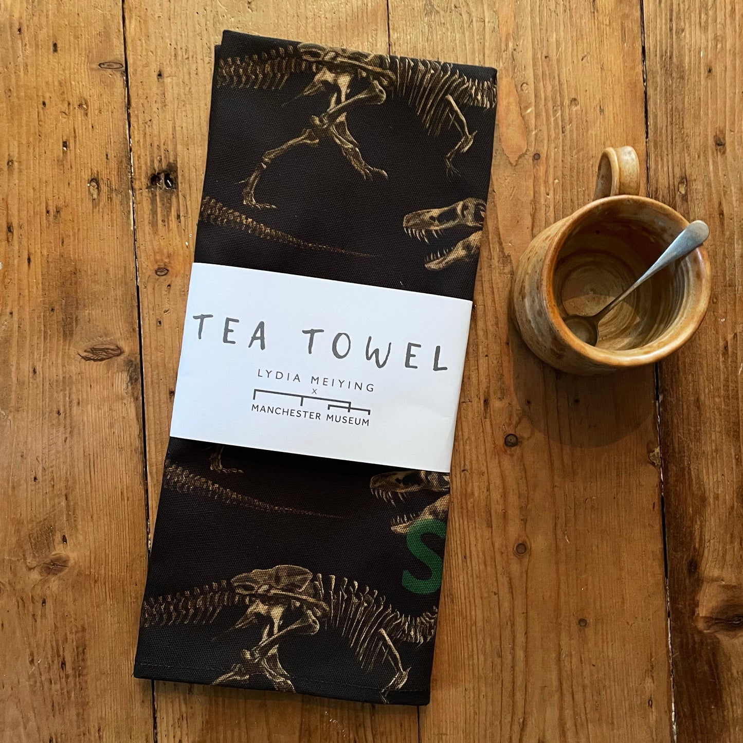 Lifestyle shot of the packaged tea towel on a rustic wooden table with an earthy coloured clay mug with a teaspoon beside it.