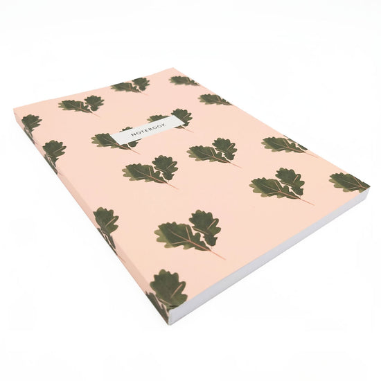 Angled side view of dusky pink notebook with pairs of illustrated acorn leaves. In a white box one thrid from the top the black text reads, notebook.