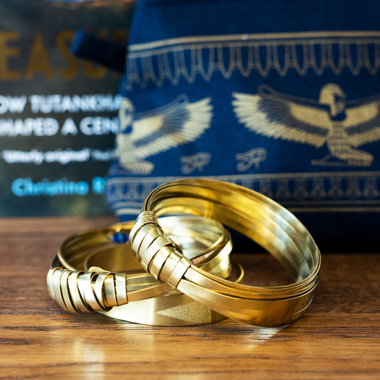 Three brass bangles, two in a ribbon style and one in a plain brass with a triangle shape embossed. An Egyptian style fabric bag is in the background.