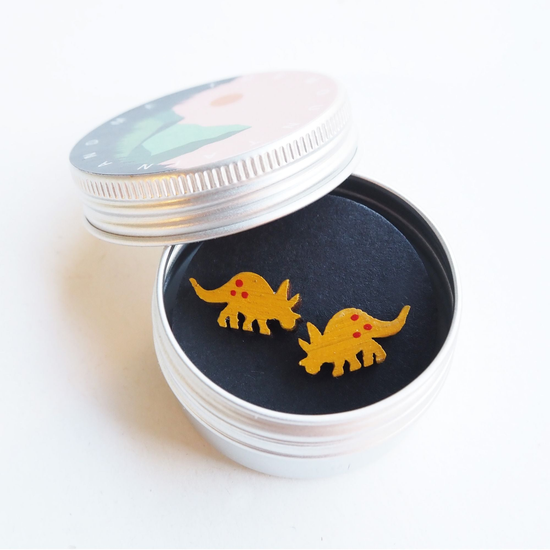 Earrings in the shape of triceratops placed in a circular metal tin against white background