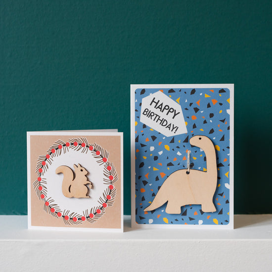 Two cards standing on a white surface with a teal background. the card on the right is smaller and square with a wood cut out squirrel on the front while the larger A6 card on the left has a wood dinosaur.