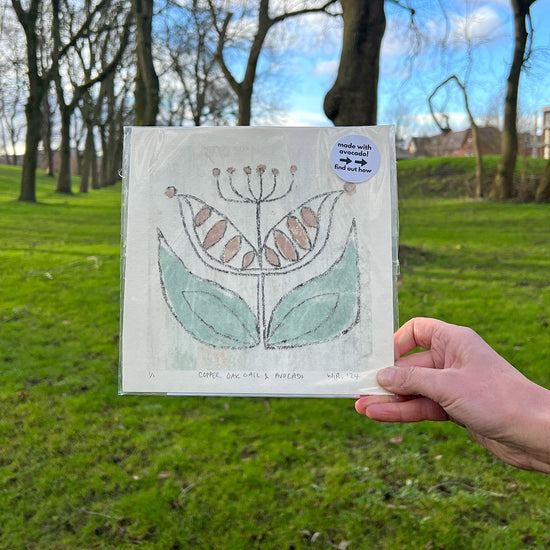 A person holding a square print with stylised flower screen printed on it in front of a park