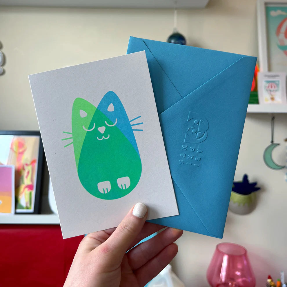 hand holding a greetings card with the blue envelope. The cat has a risograph cat print in two tones, green and blue.