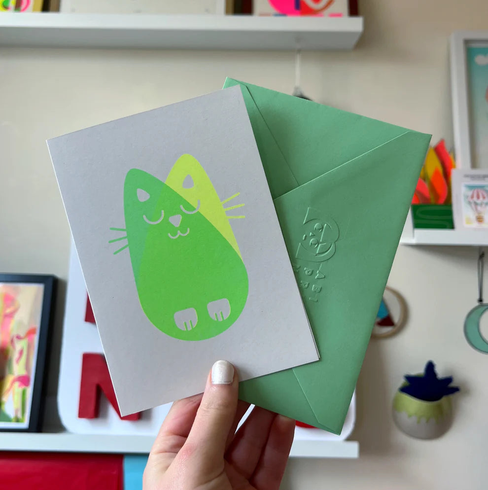 Hand holding greetings card and green envelope. The cat has a cat risograph print in two colours, green and yellow.