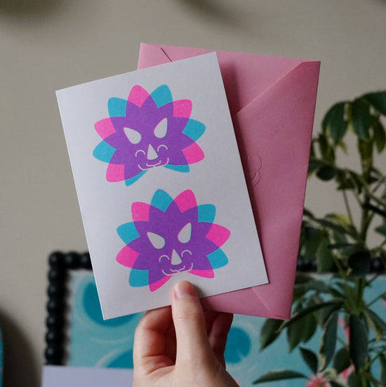 Hand holding a card with colorful Triceratops head designs and a pink envelope.