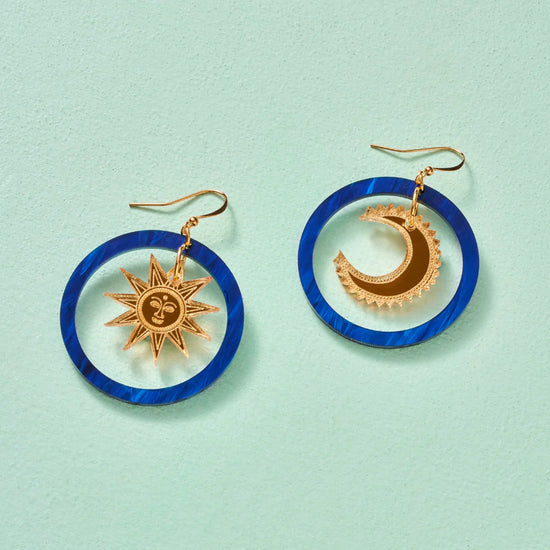 Round blue earrings with a pendant in the centre of each circle. The earring to the left has a sun with a face engraved as the pendant while the righthand earrings has a crescent moon. Gold hook fastenings. Pale sage green backdrop.