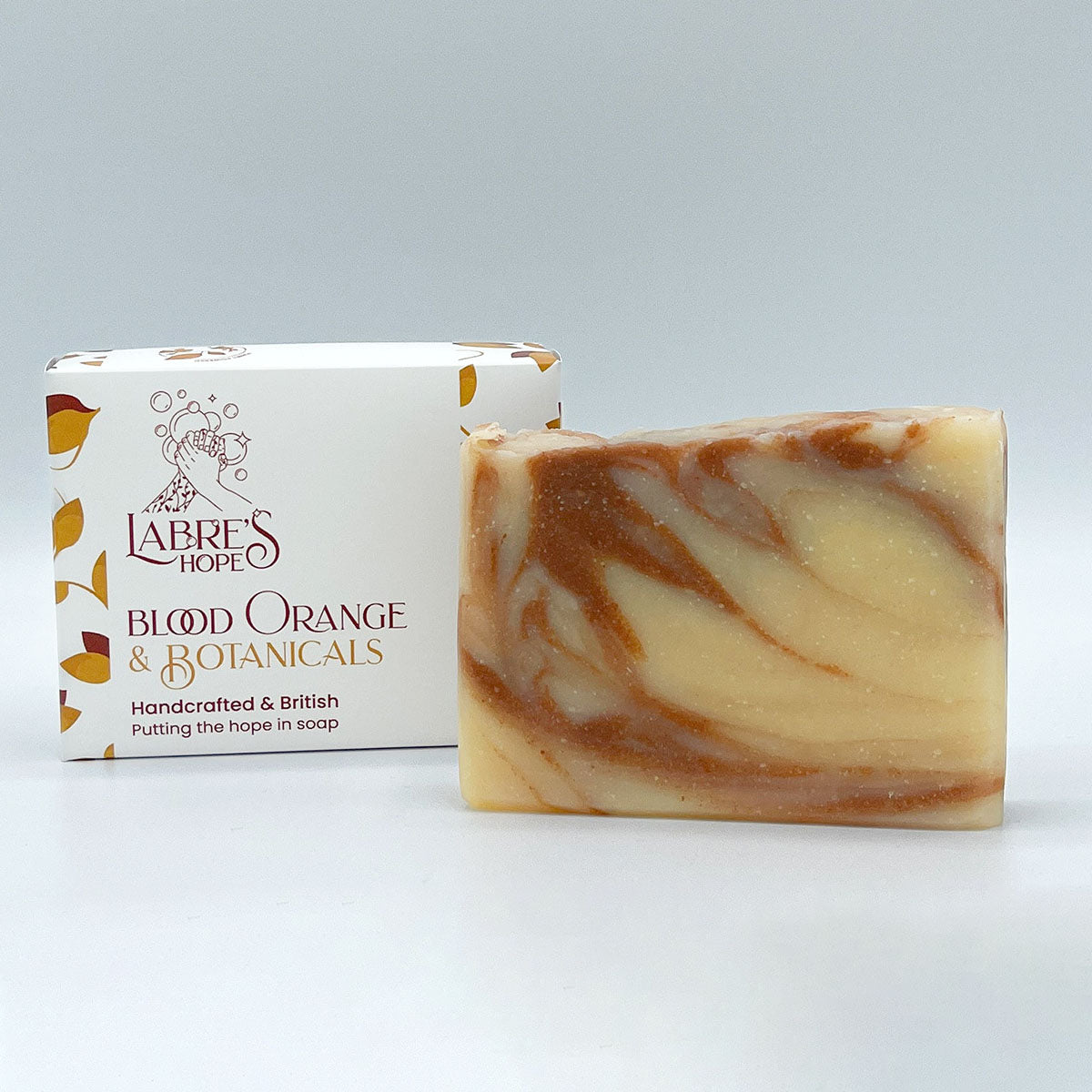 A rectangular bar of soap with a white base and swirls of orange and brown. The soap is resting inside a white cardboard box with a black and white label. 