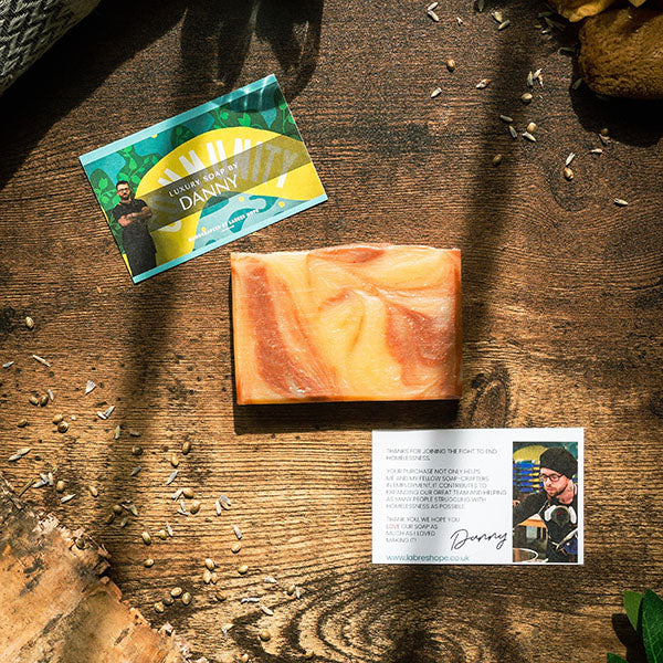  rectangular bar of soap with a white base and swirls of orange and brown. The soap is placed on a wooden surface with dried flowers and leaves scattered around it. A business card and a small note from Danny are also placed near the soap. 