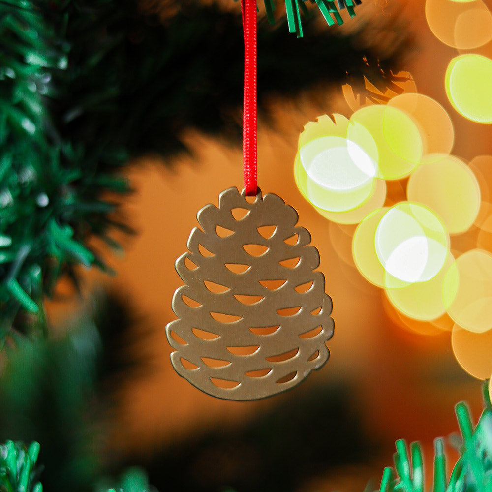 A brass pine cone Christmas ornament hanging from a red ribbon on a Christmas tree. The ornament is simple and minimalist.