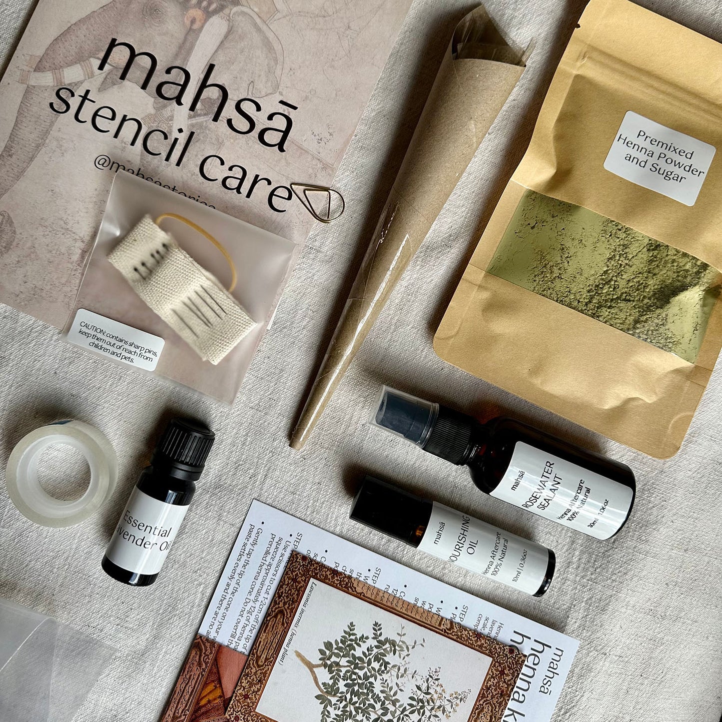 A henna kit on a white surface. The kit includes a premixed henna powder and sugar packet, a henna cone, a stencil, a needle and thread, tape, essential oils, rosewater sealant, and a henna kit guide.