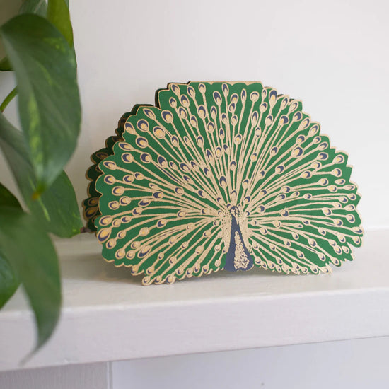 Green and gold peacock card standing upright on a white shelf with green leaves to the left.