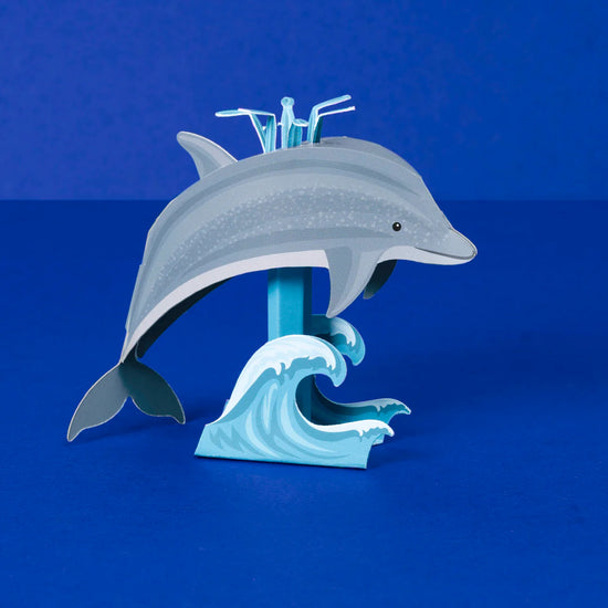 A dolphin made out of card riding a wave, placed in front of an electric blue background.