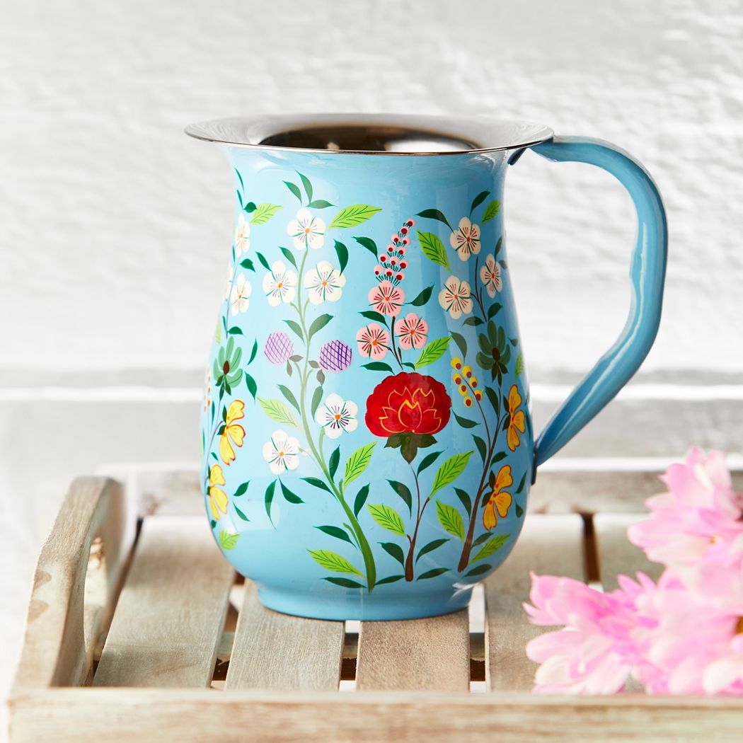 Lifestyle of a light blue enamel jug with floral patterns painted around the sides. The Jug is standing on a white washed wooden tray.