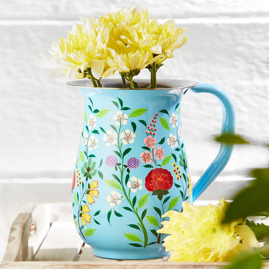 Lifestyle of a light blue enamel jug with floral patterns painted around the sides. The Jug is standing on a white washed wooden tray and has yellow flowers in it.