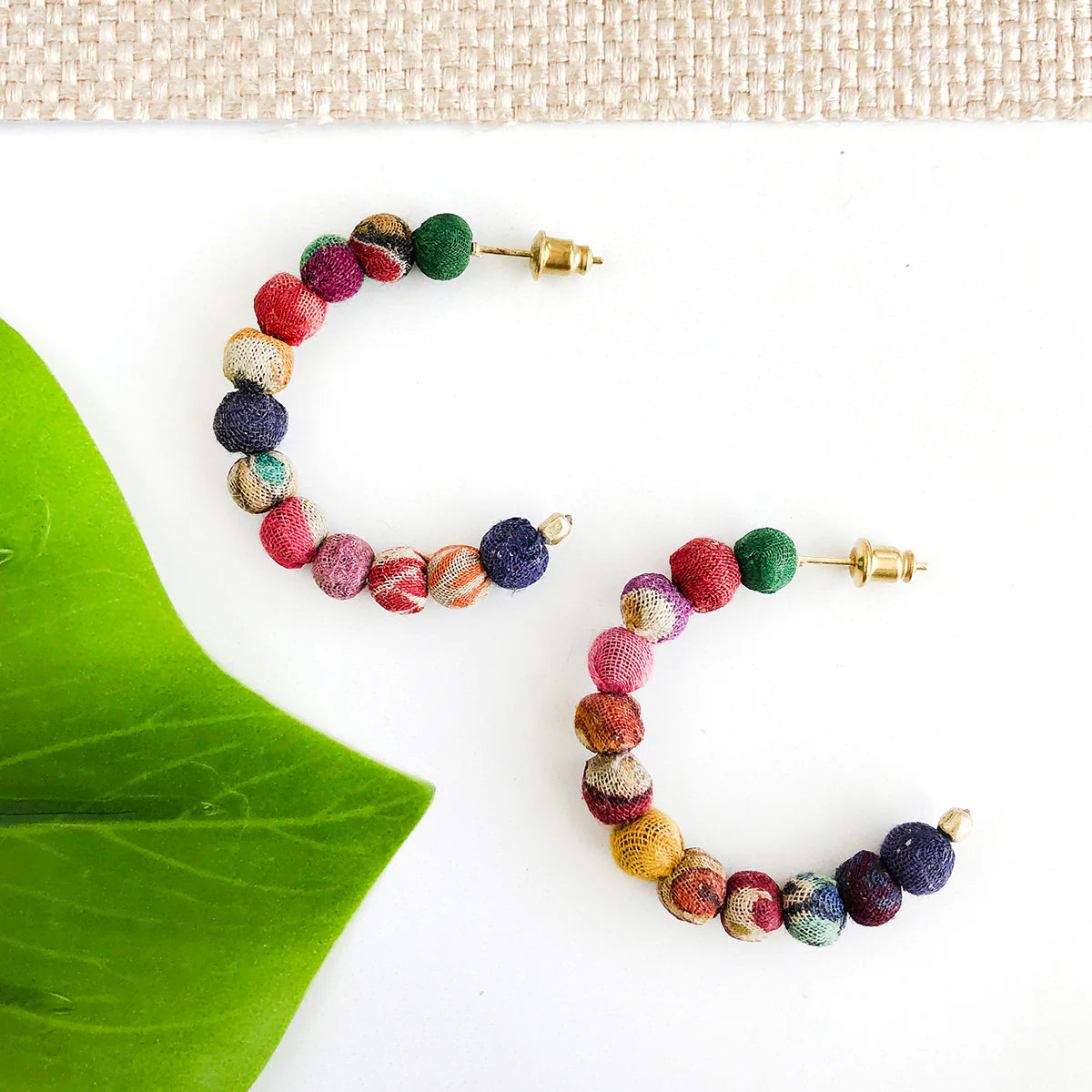 Colorful beaded half-hoop earrings on a white surface with a green leaf