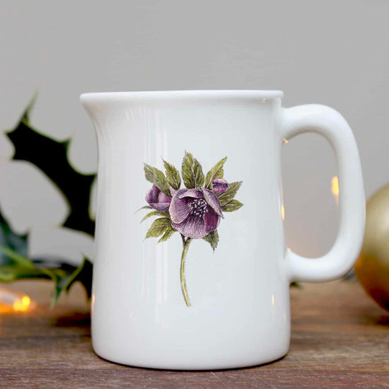 White jug with an illustration of a hellebore flower, photographed on a wooden surface with some holly in the background