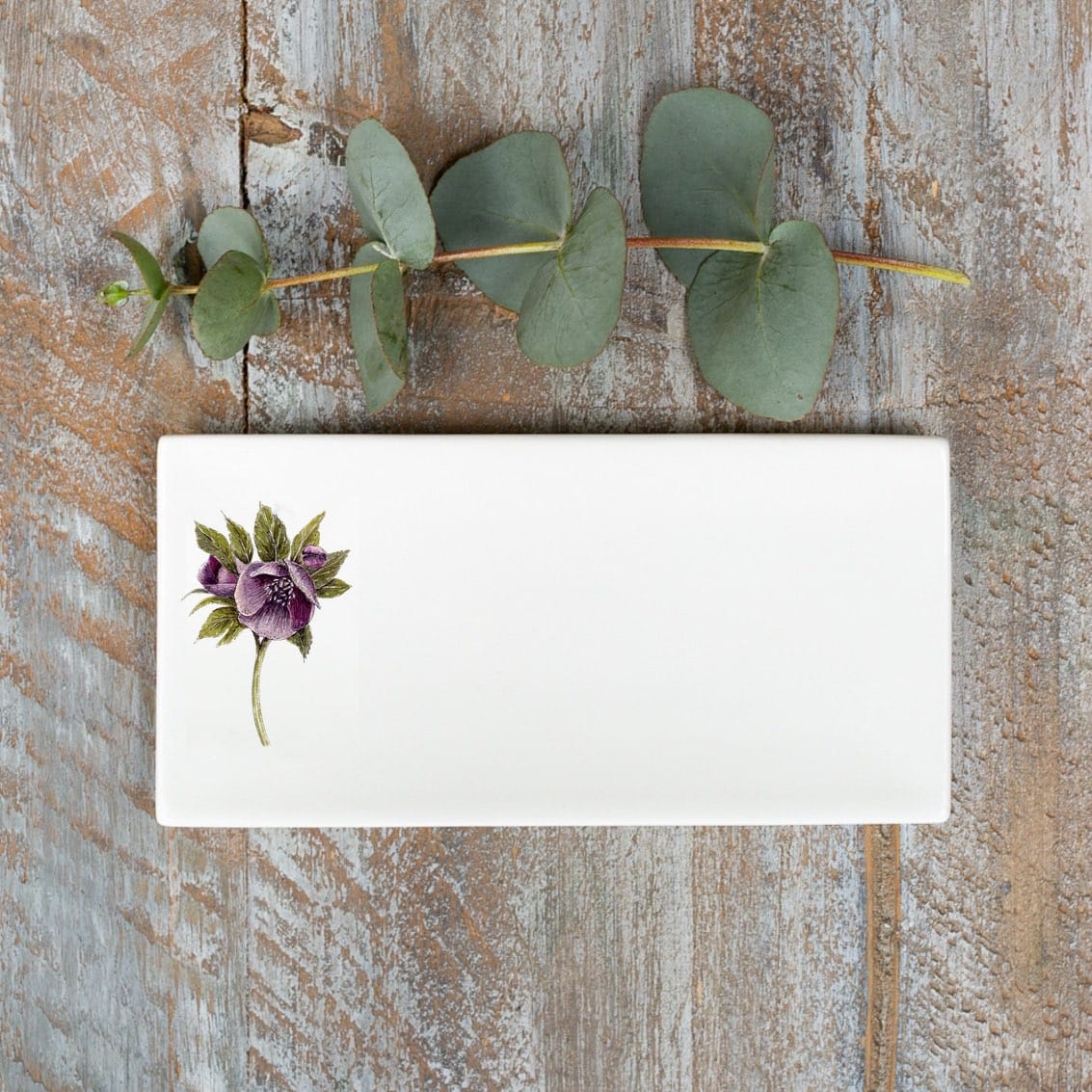White rectangular soap dish with an illustration of a hellebore flower, photographed birds-eye view on a wooden surface