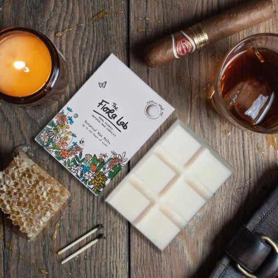 Lifestyle photograph taken birds eye view of some wax melt cubes, some wax candles, surrounded by a glass whiskey, a cigar, a leather wallet and some used matches.
