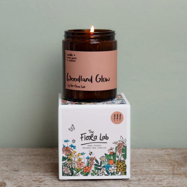 Glass candle with orange The Flora Label branded sticker, photographed on top of the illustrated packaging, on a wooden table in front of a green wall.