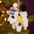 A felted wool dog Christmas ornament hanging from a green pine tree branch with warm yellow bokeh lights in the background. The ornament is white with black spots, wearing a colorful knit hat and a yellow scarf.
