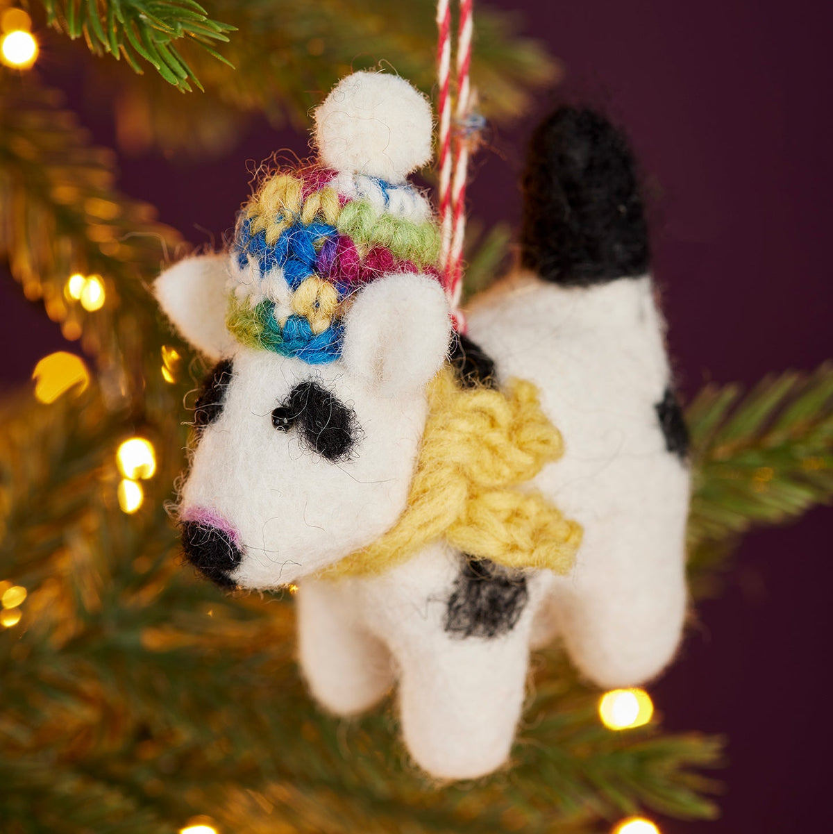 A felted wool dog Christmas ornament hanging from a green pine tree branch with warm yellow bokeh lights in the background. The ornament is white with black spots, wearing a colorful knit hat and a yellow scarf.