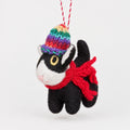 A handmade felt Christmas ornament shaped like a black and white cat wearing a colorful knit hat and a red scarf, hanging on a Christmas tree with warm lights in the background.
