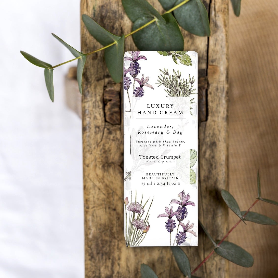 Lifestyle photo of luxury hand cream in the box it's presented in. The box is white with lavender and herb illustrations and is placed on a rustic wooden surface.