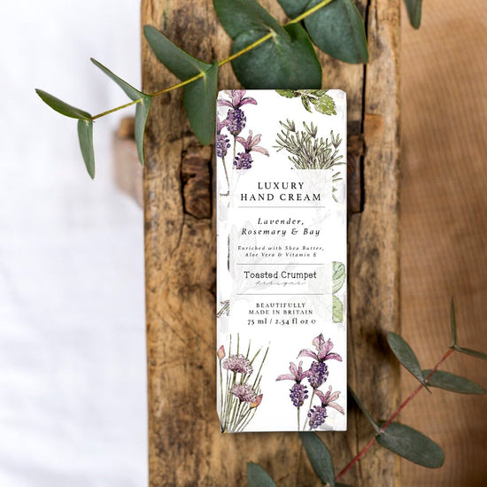 Lifestyle photo of luxury hand cream in the box it's presented in. The box is white with lavender and herb illustrations and is placed on a rustic wooden surface.