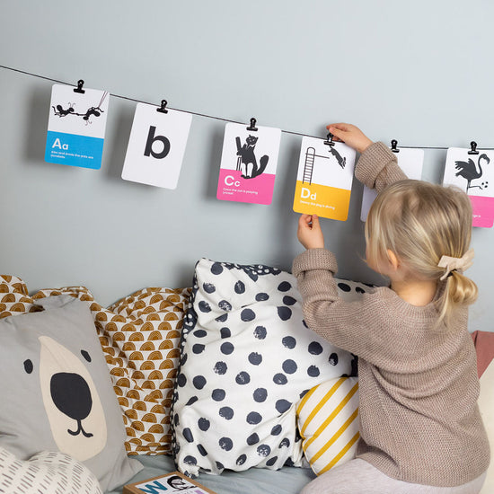 Load image into Gallery viewer, Another lifestyle with a string of phonics cards hung up by the side of a kids bed and a child is removing or hanging up a d card on the string.
