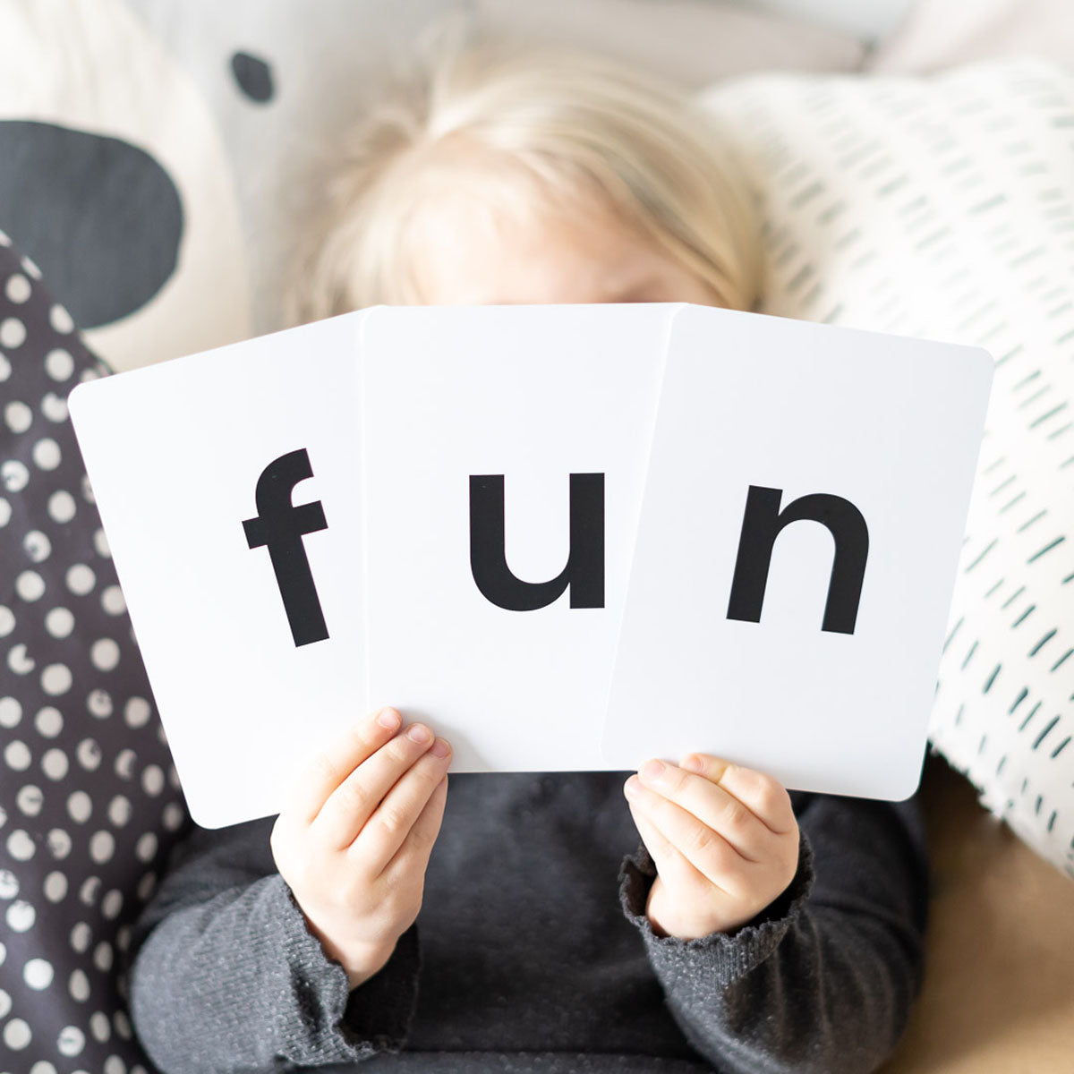 Load image into Gallery viewer, Lifestyle shot of three phonics cards onthe white and black side spelling out &amp;#39;fun&amp;#39;. The cards are held by a child in front of their face as they lie on a bed and pillows.

