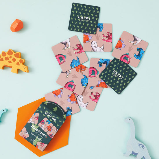 Lifestyle shot with the memory game scattered in front of an angled game pack and lying on a pale turquoise surface. Wooden dinosaurs can bee seen lying around the game as well.