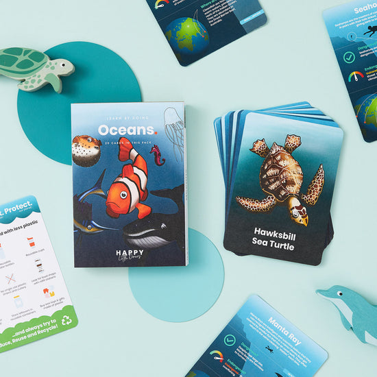 Load image into Gallery viewer, Lifestyle shot of the card pack from the front and a stack of the cards to the right of th epack. A wooden dolphin and turtle can be seen peeking from the right and left side of the shot respectively.
